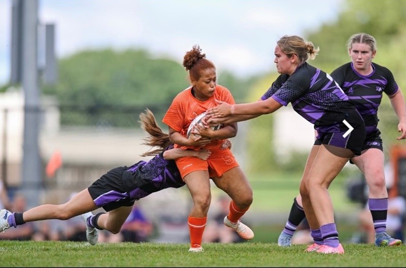 Mariah Carter holding the ball and breaking a tackle in a rugby match.