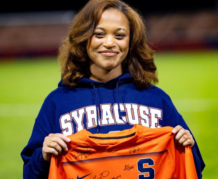 Maria carter smiling in a Syracuse hoodie holds up an orange, signed Syracuse jersey on a stadium field for hometown hero.